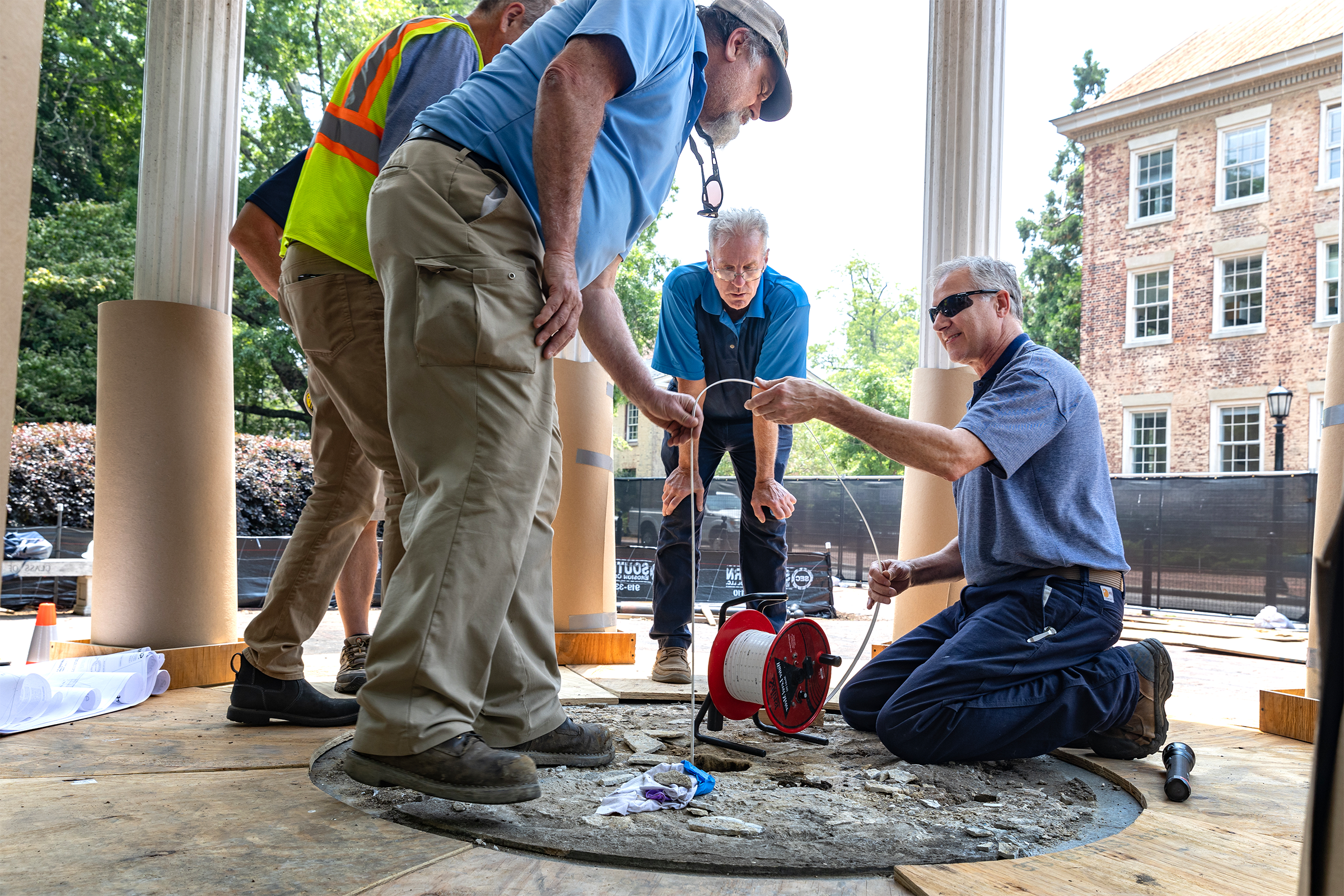 Four workers, one kneeling and the other three standing, working to measure the depth of a well.