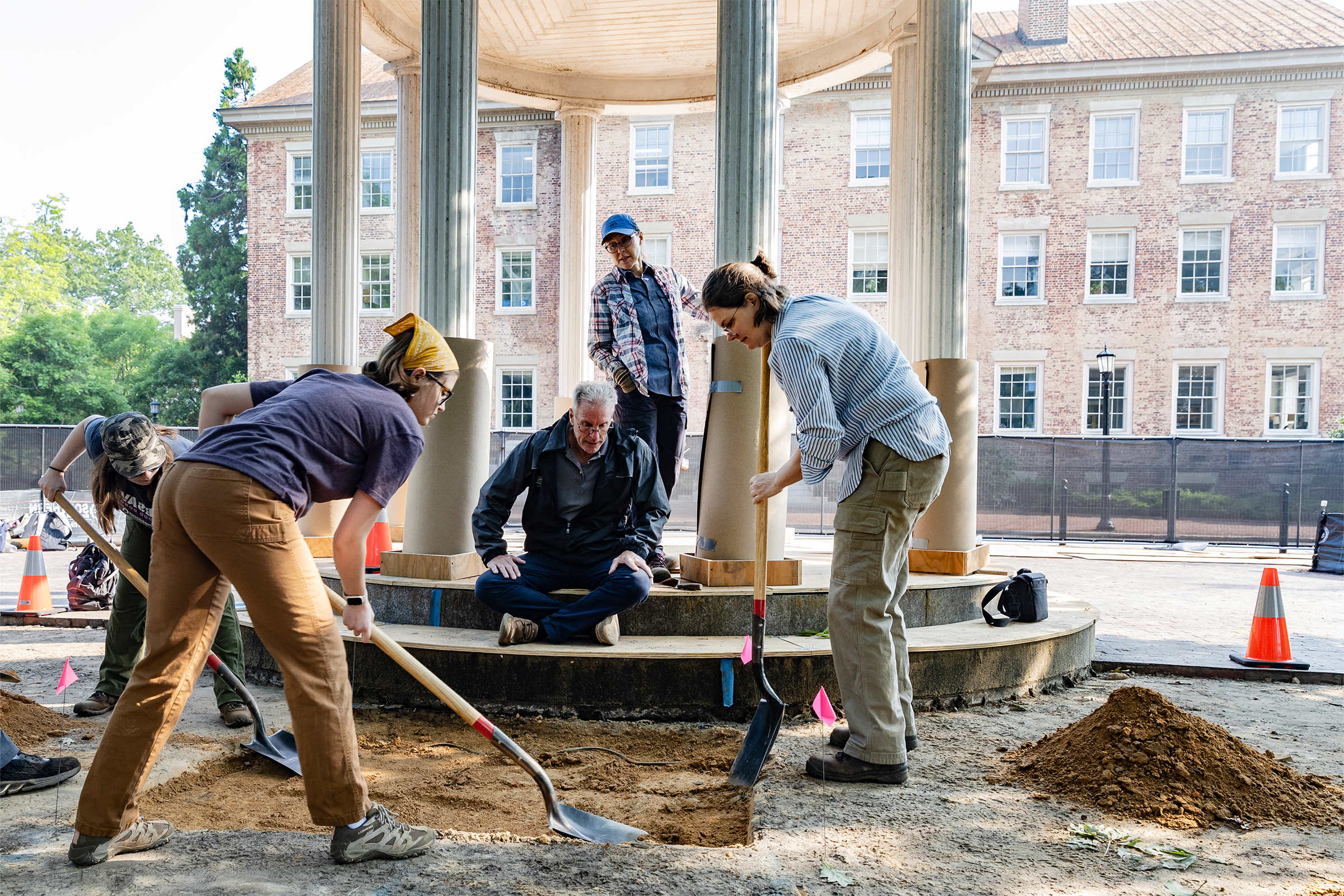 Two students using shovels to dig into the ground next to a well. Two onlookers are in the background.