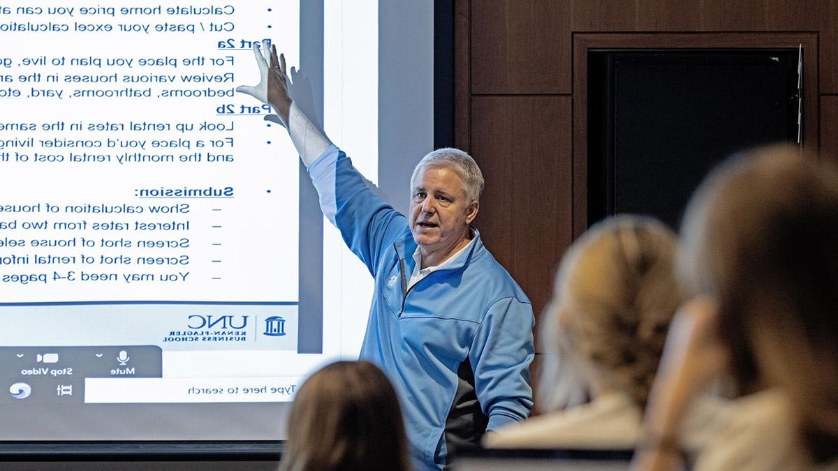 Chip Snively Jr. wearing Carolina Blue shirt and pointing to text projected on screen in front of classroom.